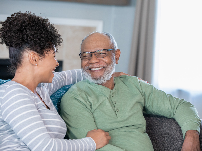 A senior citizen and his daughter smile at one another on the couch of his assisted living apartment.