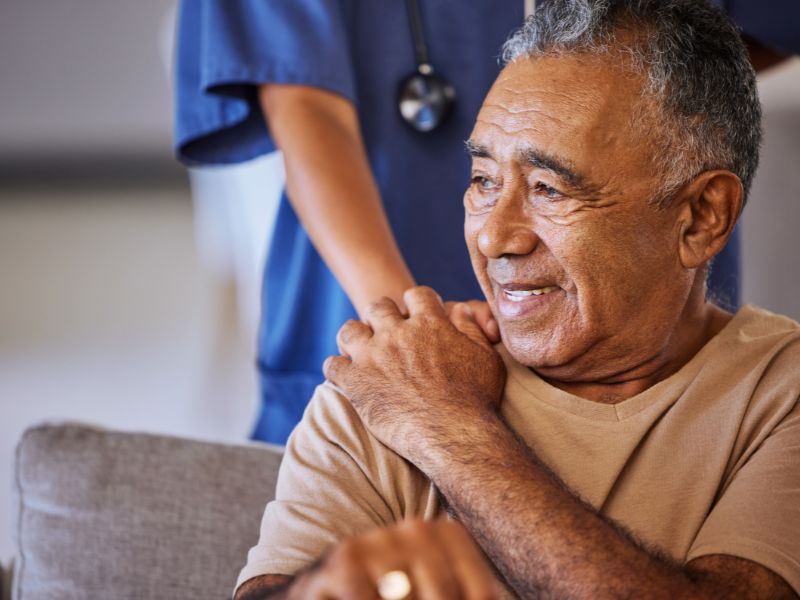 A senior smiles as he rests his hand upon the hand of a caregiver, who has placed her hand on his shoulder at an assisted living facility