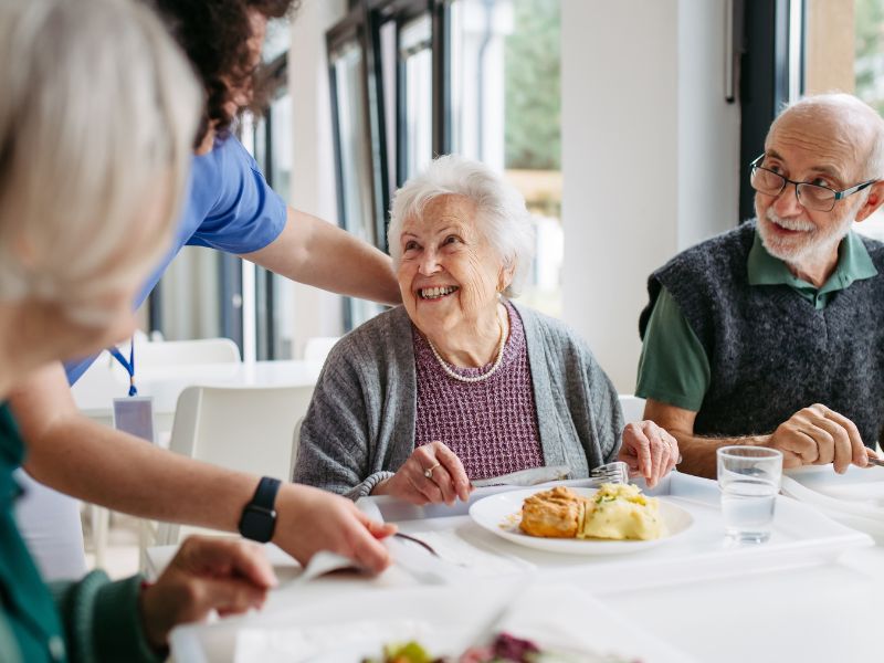 A group of elderly people smile at a caregiver at a senior living facility